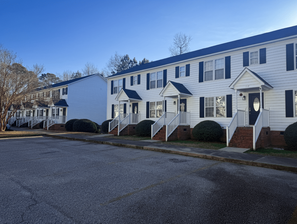 Row of white two-story townhouses with black shutters and small front porches, set alongside a paved parking area on a clear day.